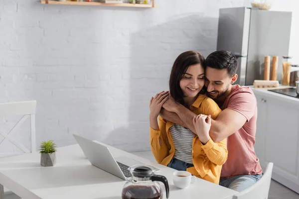 Sonriente Joven Abrazando Novia Con Ordenador Portátil Cocina —  Fotos de Stock