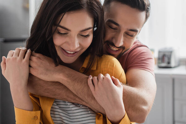smiling young man with closed eyes hugging girlfriend at home