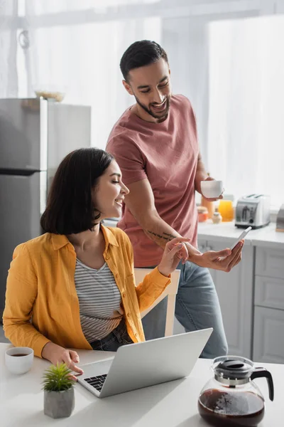 Sorrindo Jovem Mostrando Celular Para Namorada Sentado Com Laptop Casa — Fotografia de Stock