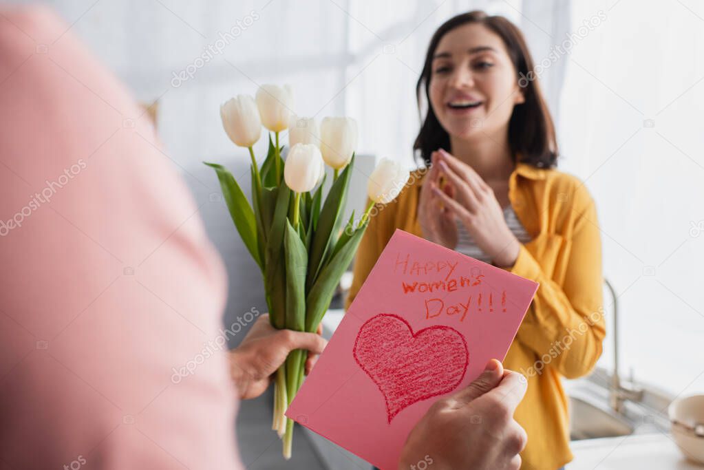 young man holding bouquet of flowers and greeting card near blurred girlfriend in kitchen