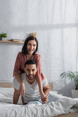 Yatakta beyaz çarşaflarla oturan gülümseyen genç çift, kameraya bakıyor ve yatak odasında sarılıyorlar.