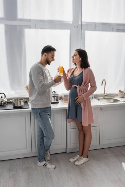 笑着站在孕妇身边的年轻人在厨房里喝着橙汁 — 图库照片