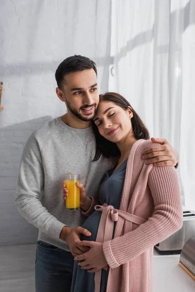 Sorridente Jovem Abraçando Mulher Grávida Com Suco Laranja Cozinha — Fotografia de Stock