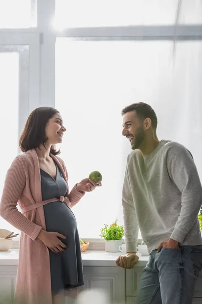 一个在厨房里拿着苹果站在孕妇身边的积极的年轻人 — 图库照片