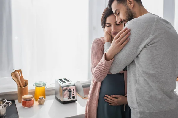Smiling Young Man Closed Eyes Hugging Pregnant Woman Kitchen — 스톡 사진