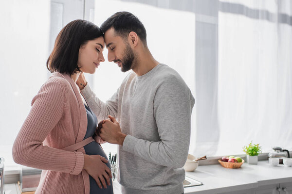 young man with closed eyes gently holding hand of pregnant woman in kitchen