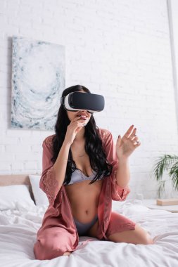 VR kulaklıklı, ipek bornozlu ve yatakta oturan seksi iç çamaşırlı mutlu genç bir kadın.