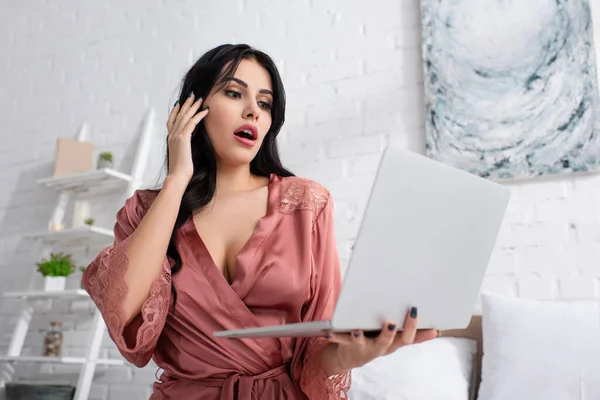shocked young woman in silk robe looking at laptop