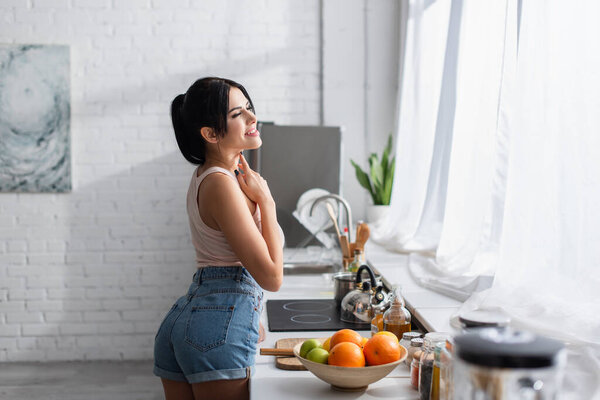 cheerful young woman standing near bowl with fresh fruits in kitchen 