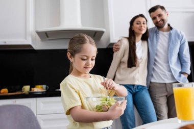 Bulanık arka planda mutlu ebeveynlerin yanında sebze salatası karıştıran kız.