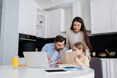 Babasıyla ödev yaparken kızının yanında gülümseyen bir kadın kitap okuyor.