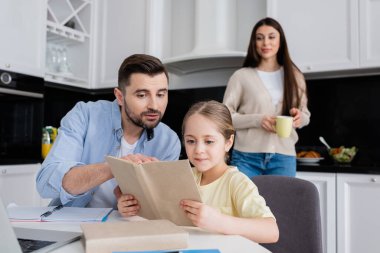 Adamın biri, kızı ile ödev yaparken kitabı işaret ediyor bulanık arka planda gülümsüyor.