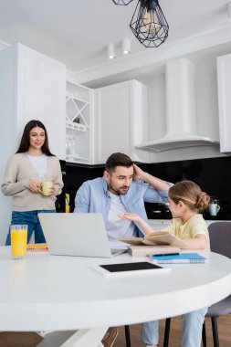 Annesi gülümserken, babasıyla ev ödevi yaparken elini gösteren kız.