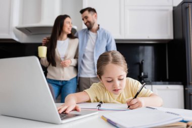child using laptop and writing in notebook near smiling parents talking on blurred background clipart