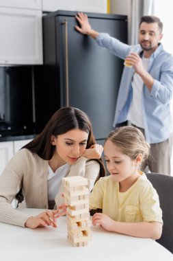 Anne ve kızı odun blokları oynuyorlar. Bulanık bir babanın yanında. Mutfakta portakal suyu içiyorlar.