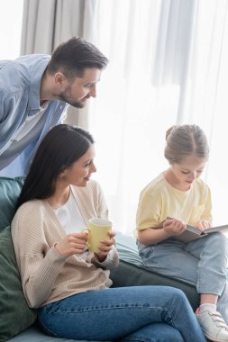 Anne ve babanın yanında bir fincan çayla kitap okuyan kız.
