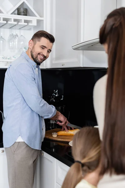 Joyful Man Cutting Bread Blurred Wife Daughter Hugging Kitchen — Stock Photo, Image