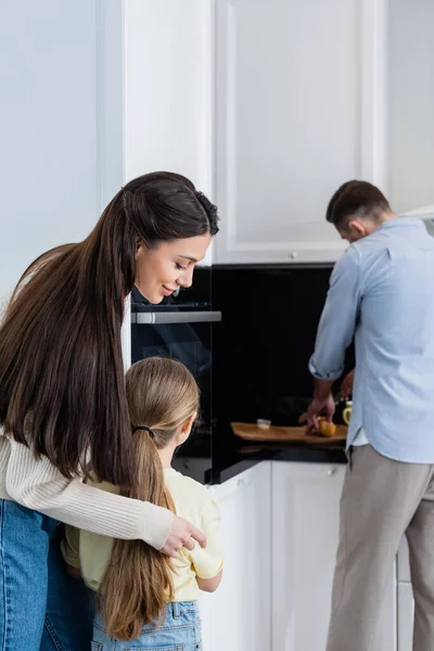 Leende Kvinna Omfamna Dotter Nära Suddig Man Skära Bröd Köket — Stockfoto