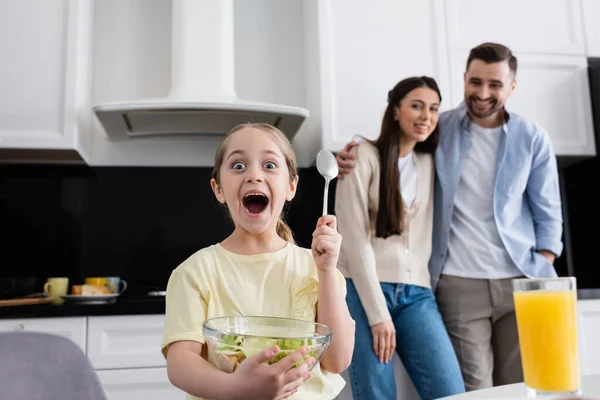 Upphetsad Flicka Innehav Sked Och Grönsak Sallad Nära Föräldrar Ler — Stockfoto