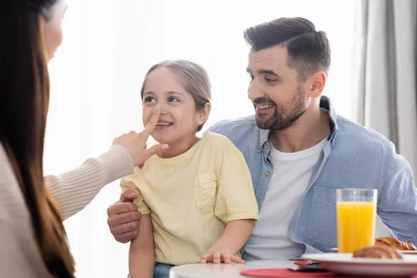 朝食中に幸せな夫の近くの明るい娘の鼻に触れる母親 — ストック写真