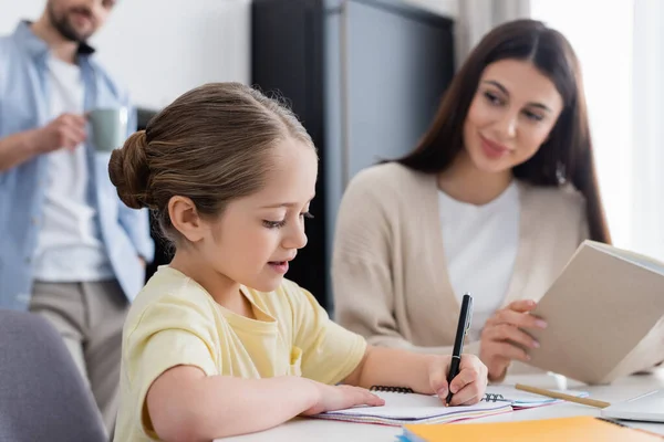 Chica Escribiendo Cuaderno Cerca Borrosa Mamá Papá Con Taza Café — Foto de Stock