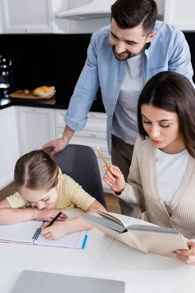 Sonriente Hombre Pie Cerca Esposa Leyendo Libro Cerca Hija Escribiendo —  Fotos de Stock