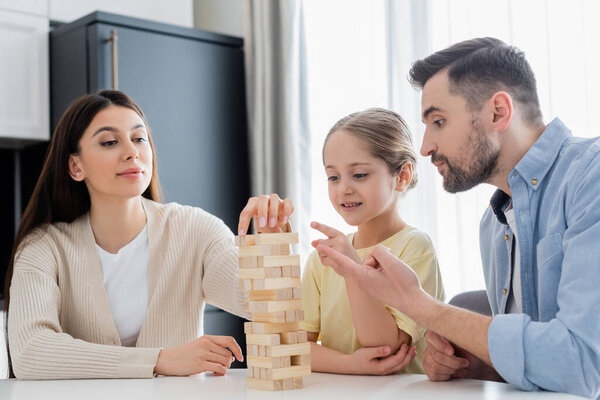 father and daughter pointing with fingers while mother removing block from wooden tower