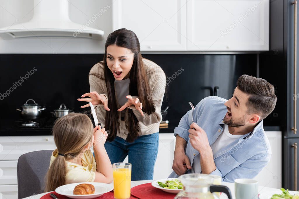 woman having fun while scaring daughter near excited husband during breakfast