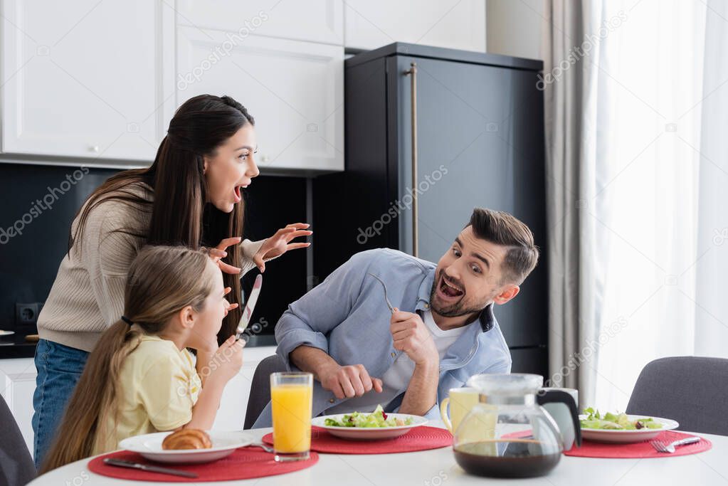 woman scaring excited husband and daughter having breakfast in kitchen