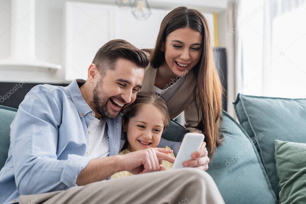 laughing man pointing on mobile phone near happy daughter and wife at home