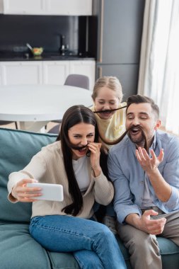 Neşeli aile akıllı telefondan selfie çekerken anne saçlarıyla bıyık taklidi yapıyor.