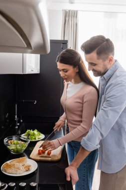 Gülümseyen kadın mutlu kocaya salata hazırlarken kiraz domatesleri kesiyor.