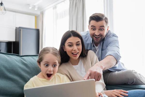 cheerful man pointing at laptop near excited wife and daughter