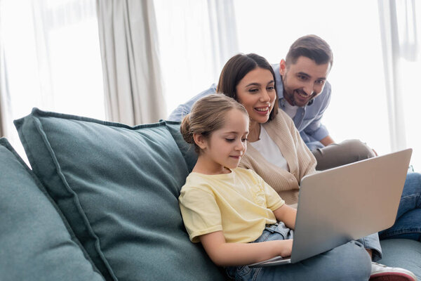 happy parents looking at daughter using laptop on couch at home
