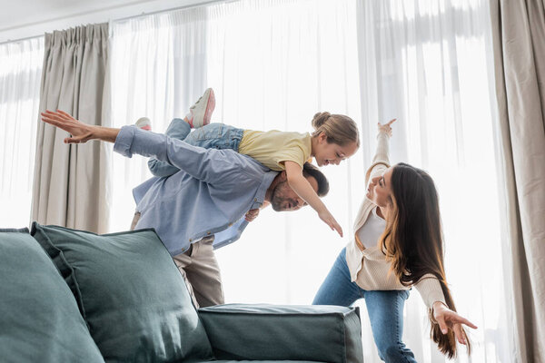 joyful girl piggybacking on back of father and imitating plane with mom