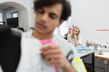young dressmaker sitting near cloth sample and sewing machine while designer working on blurred foreground clipart