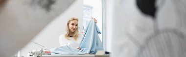 pleased fashion designer looking at tissue at workplace on blurred foreground, banner clipart