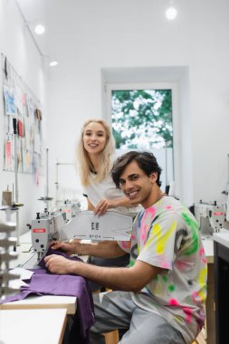 Genç moda tasarımcısı ve terzi atölyedeki dikiş makinelerinin yanındaki kameraya gülümsüyor.