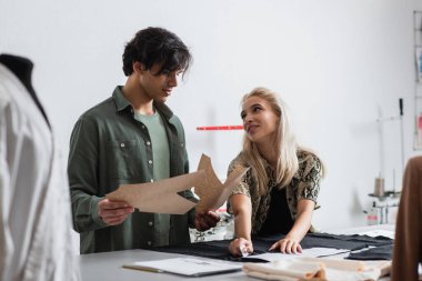 Moda tasarımcısı kumaş kesen sarışın terzinin yanında dikiş desenleri tutuyor.