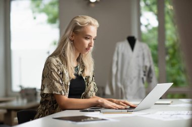 İş yerindeki cihazların yanında dizüstü bilgisayarda yazan sarışın moda tasarımcısı.