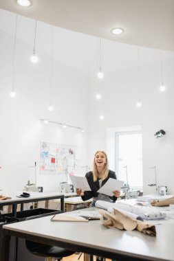 Geniş bir terzi dükkanında çalışırken heyecanlı moda tasarımcısı gülüyor.
