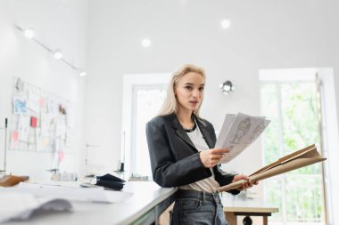 Moda tasarımcısı kameraya bakarken dikiş desenleri ve çizimler tutuyor.