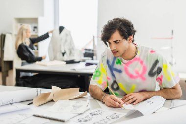 Terzi dükkanında bulanık bir meslektaş çalışırken kalemle çizimlere bakan yaratıcı moda tasarımcısı