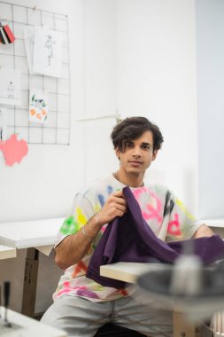 young designer looking at camera while holding sample of fabric in fashion atelier clipart
