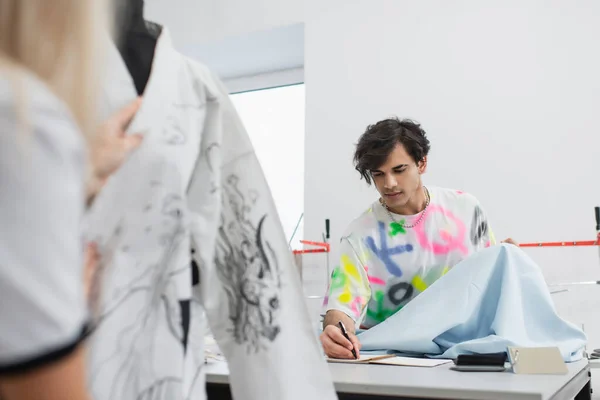 Fashion Designer Holding Tissue Sample Writing Notebook Blurred Colleague — Stock Photo, Image