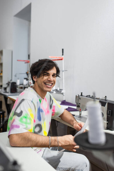happy tailor smiling at camera at workplace near sewing machine, blurred foreground
