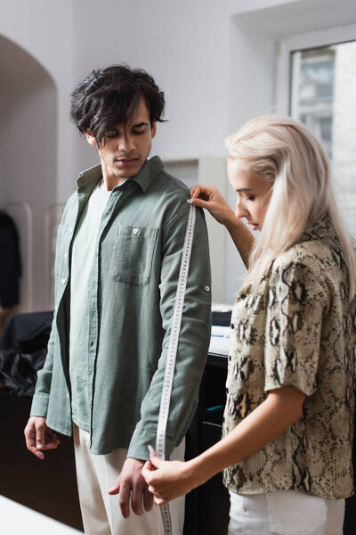 young man near blonde designer measuring him in atelier