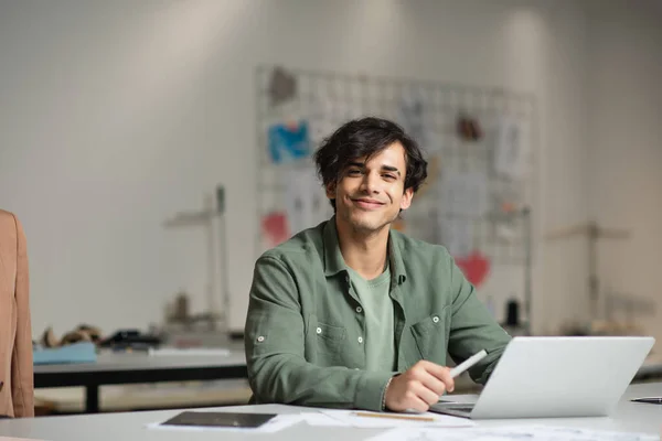 Χαρούμενος Σχεδιαστής Χαμογελώντας Στην Κάμερα Κοντά Στο Laptop Στο Εργαστήριο — Φωτογραφία Αρχείου