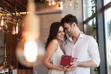joyful man holding red gift box near smiling girlfriend in slip dress in restaurant  clipart