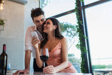 happy couple in wireless earphones looking at each other near smartphone and bottle of wine clipart